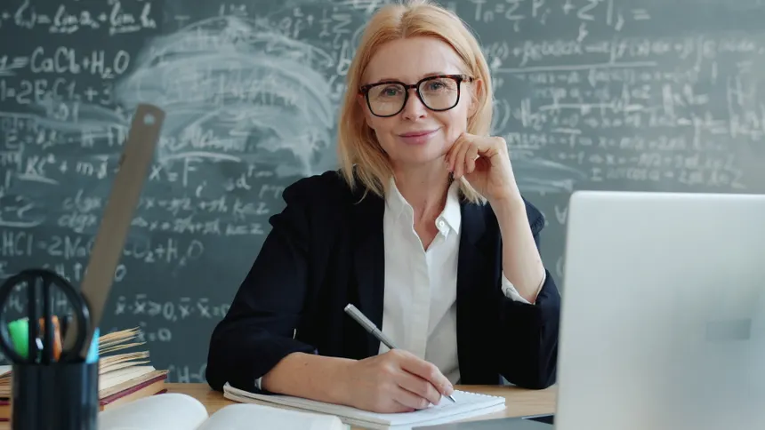 Female teacher smiles at desk with chalkboard in educational setting.