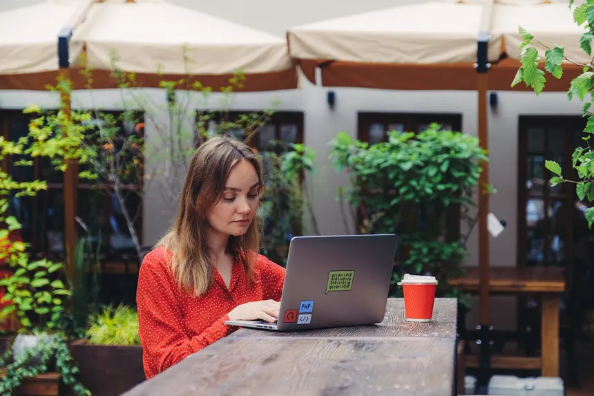 Young woman working on a laptop in an outdoor cafe setting, enjoying a cup of coffee.