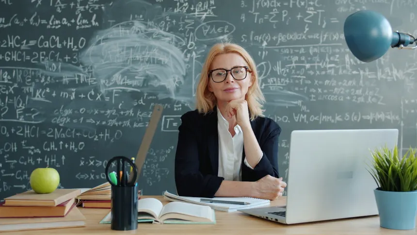 Experienced female teacher sitting in front of chalkboard with mathematical formulas.