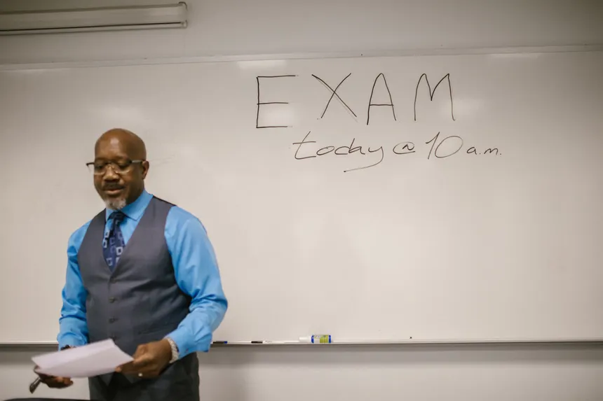 A professor stands next to a whiteboard with 'Exam today at 10 a.m.' written, indicating an upcoming exam in a classroom.